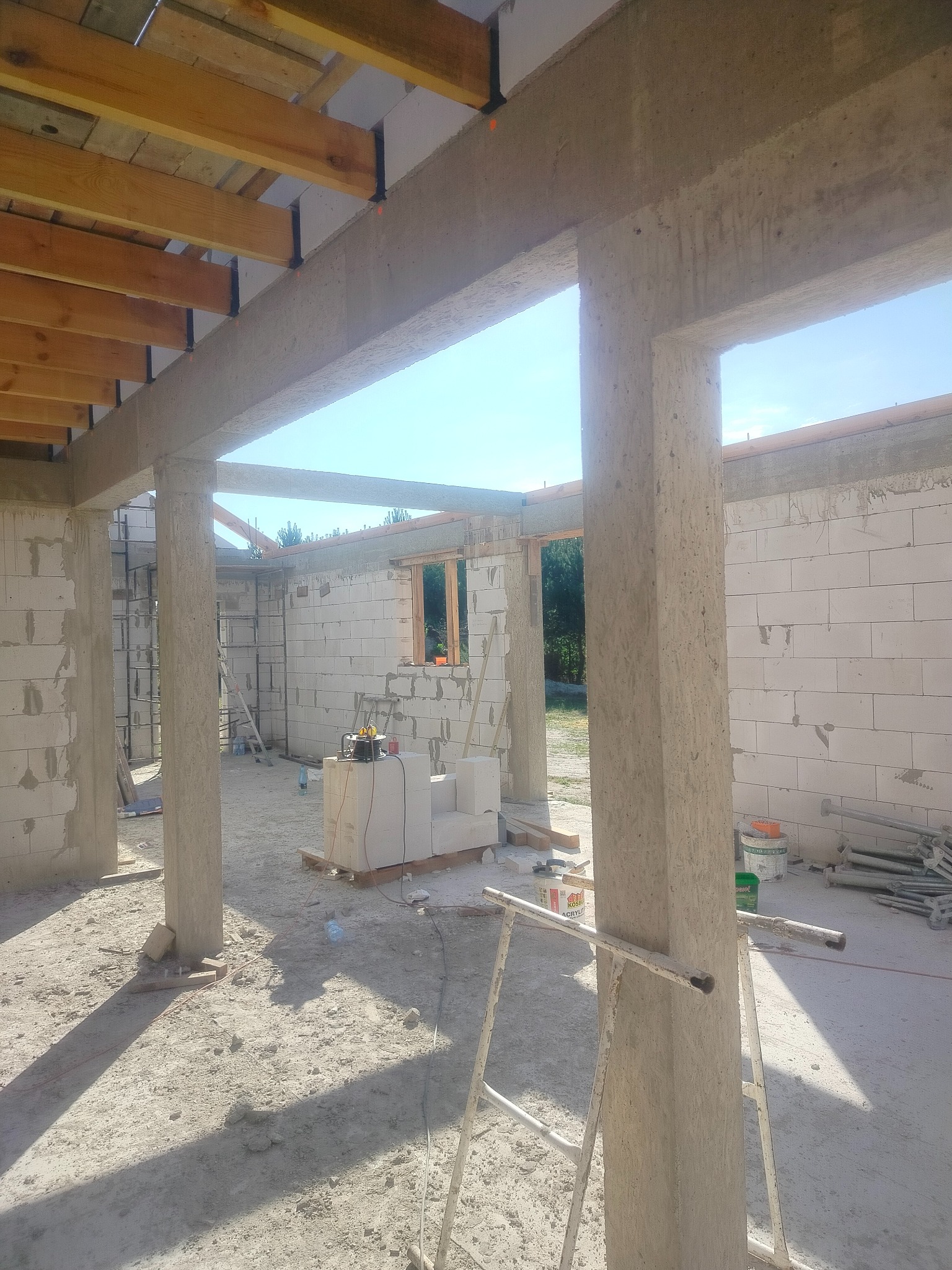 Surowy stan budynku z betonu komórkowego w trakcie budowy. Widoczne betonowe słupy, drewniane belki stropowe i częściowo wymurowane ściany. Na podłodze gruz i narzędzia.
