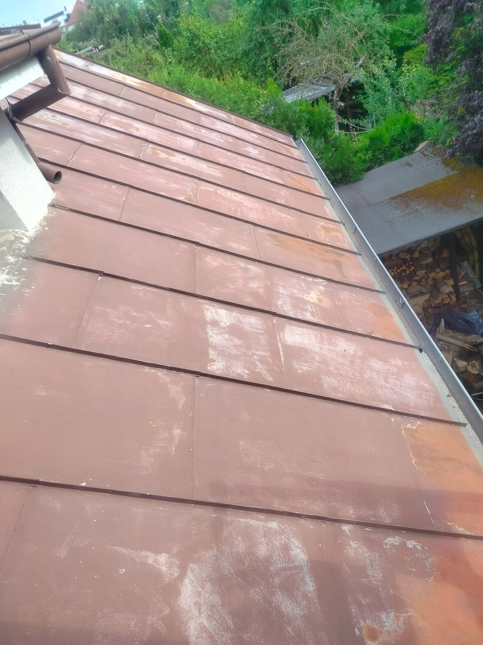 Metalowy dach z widocznymi śladami korozji i odbarwieniami, z rynną i fragmentem komina, na tle zieleni drzew i krzewów. Widok z góry, ukośny kadr.