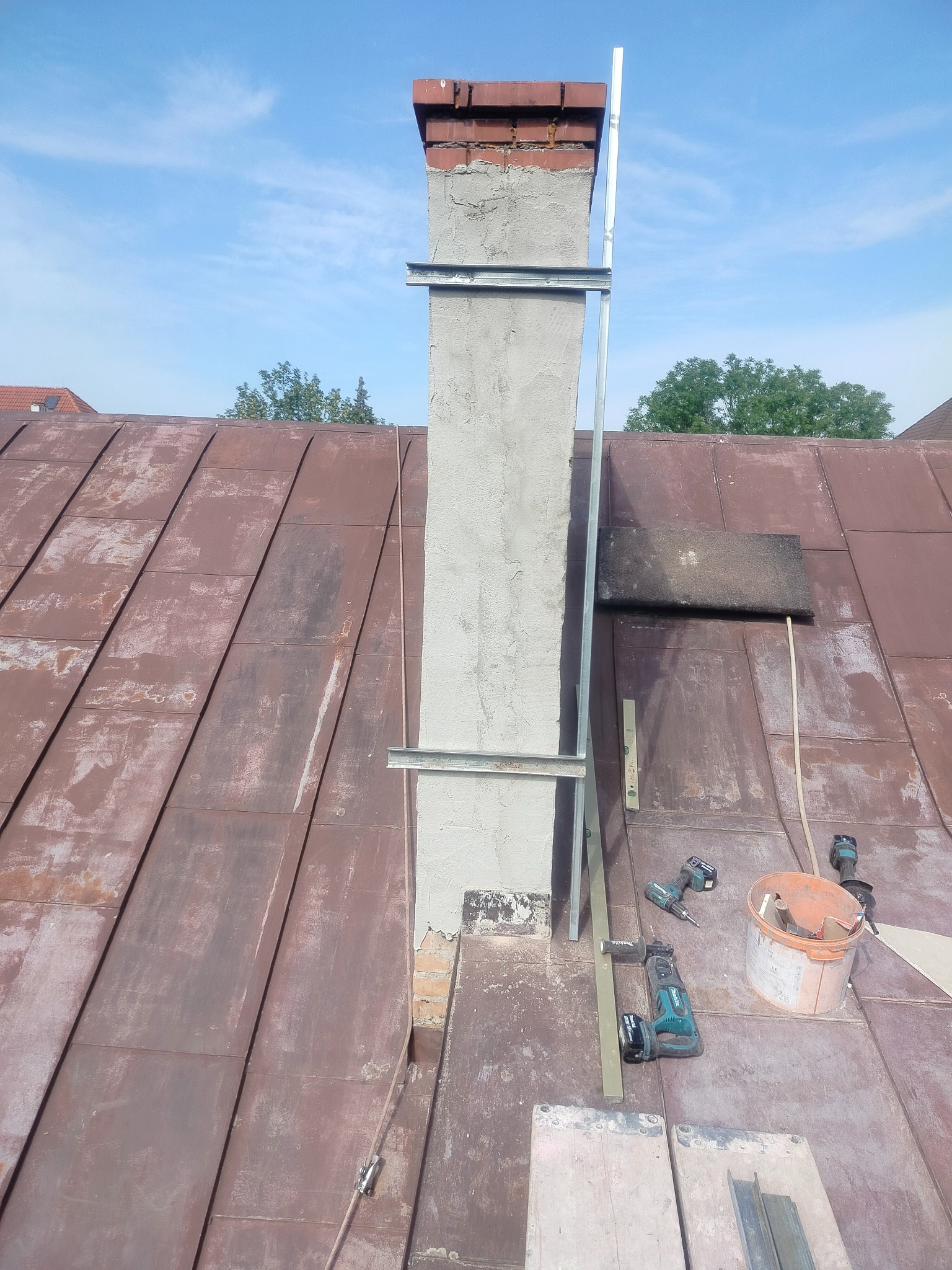 Odnowiony komin na dachu z blachy, z widocznymi narzędziami i poziomnicą. Częściowo otynkowany, z zamontowanymi metalowymi wspornikami. Błękitne niebo w tle.