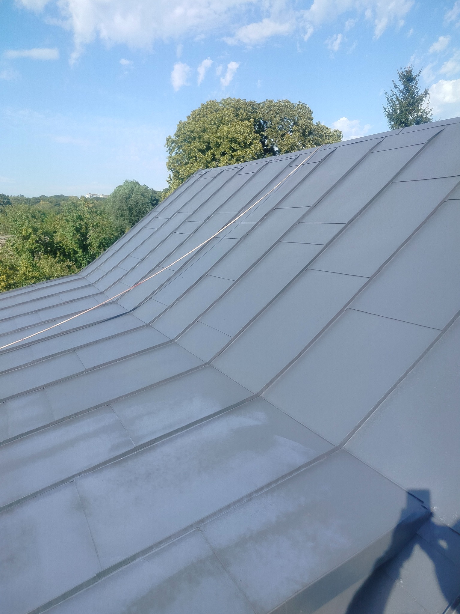 Szary, metalowy dach z widocznymi łączeniami paneli na tle błękitnego nieba i zieleni drzew. Widok z góry, ukośny kadr.