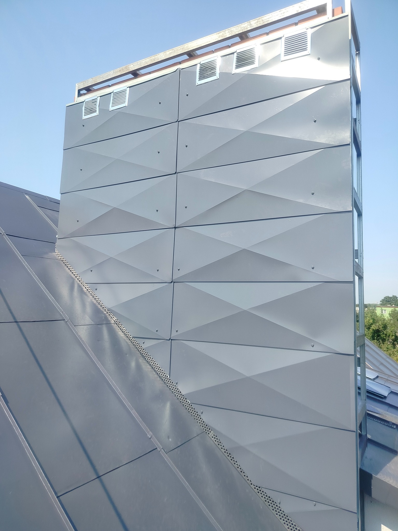 Szara, nowoczesna elewacja budynku z geometrycznym wzorem paneli, widoczna z dołu, z fragmentem dachu i elementami wentylacyjnymi. Jasne, błękitne niebo w tle.