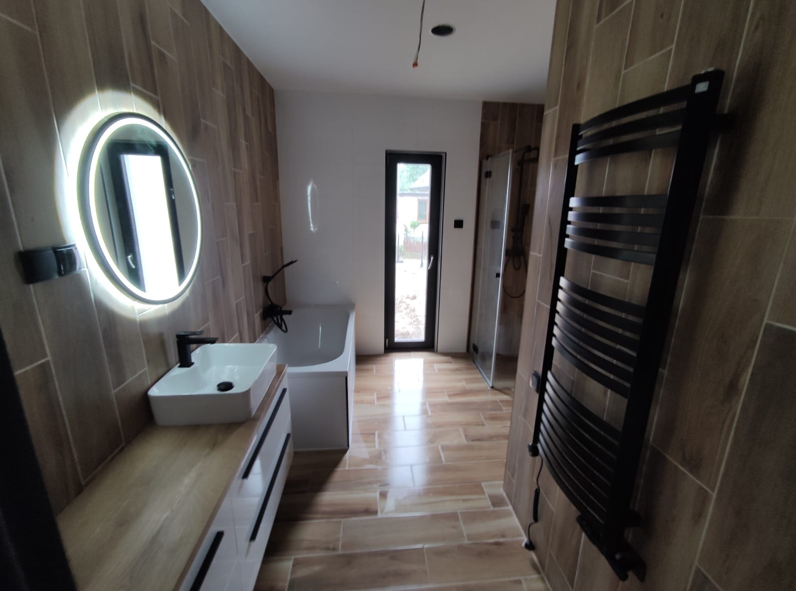 Nowoczesna łazienka z drewnopodobnymi płytkami, okrągłym lustrem z podświetleniem LED, czarną armaturą i grzejnikiem drabinkowym. Widoczna kabina prysznicowa i okno.