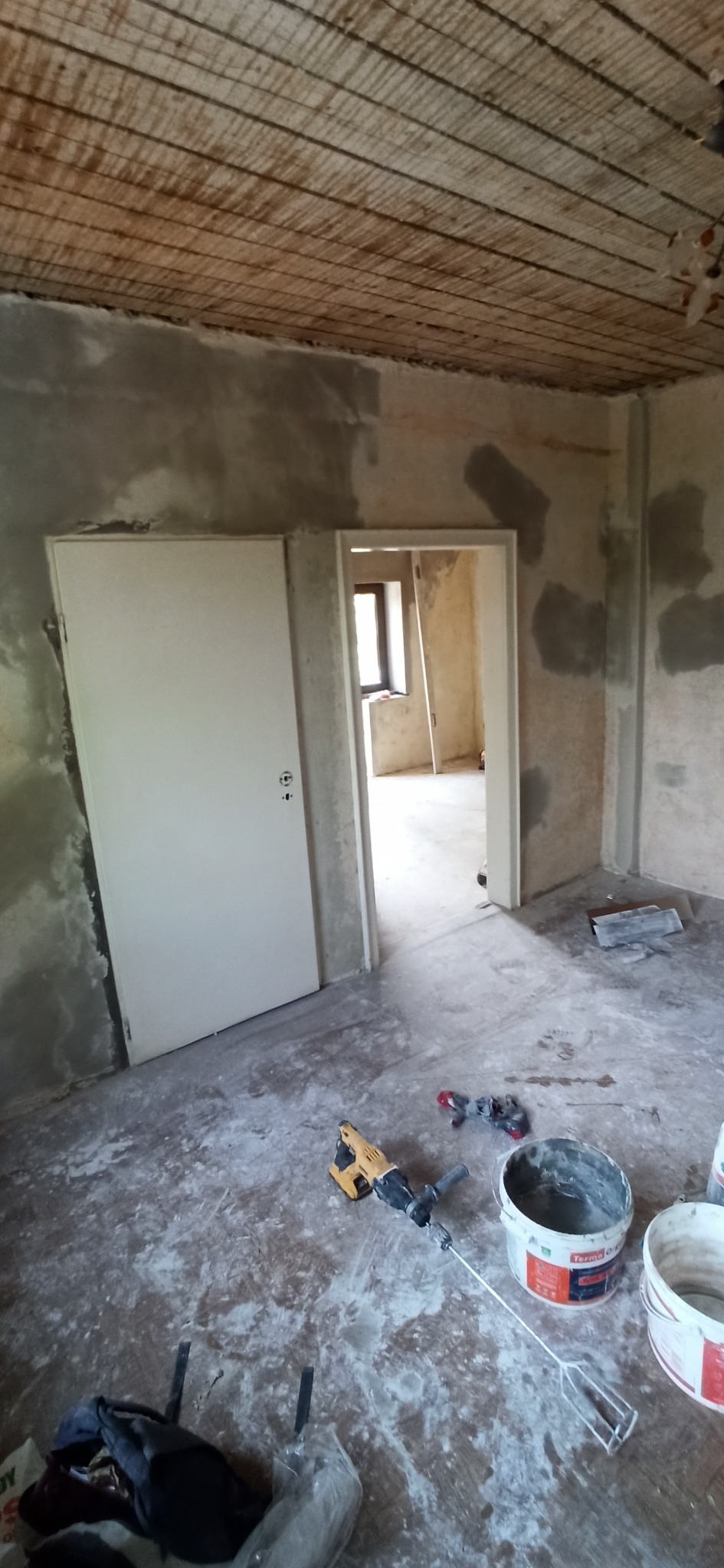 Wnętrze w trakcie remontu: surowe ściany, betonowy strop, otwarte drzwi, narzędzia do szpachlowania i mieszania zaprawy na podłodze. Widok na sąsiednie pomieszczenie.