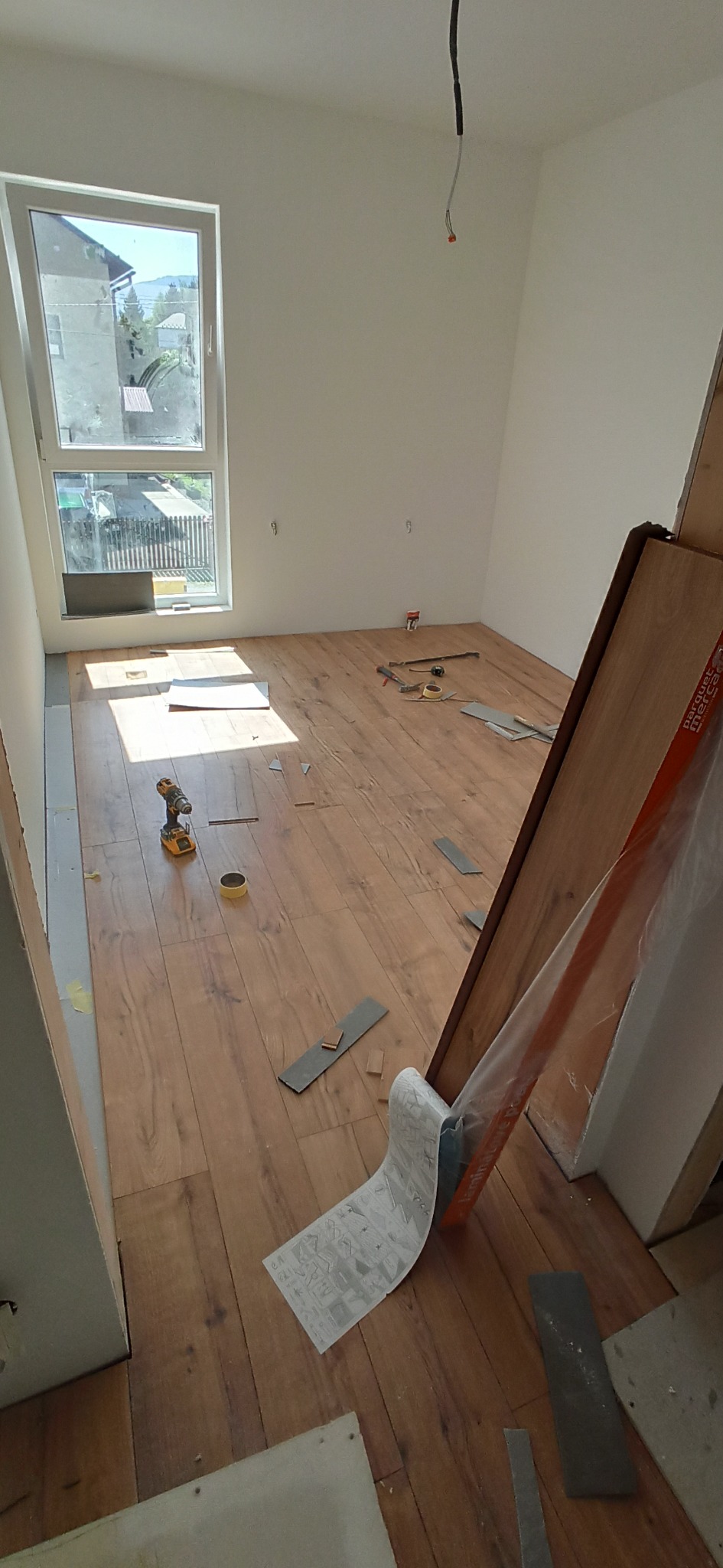 Układanie paneli podłogowych w jasnym pokoju z oknem. Widoczne narzędzia, panele i częściowo ułożona podłoga. Słoneczne światło wpada przez okno.