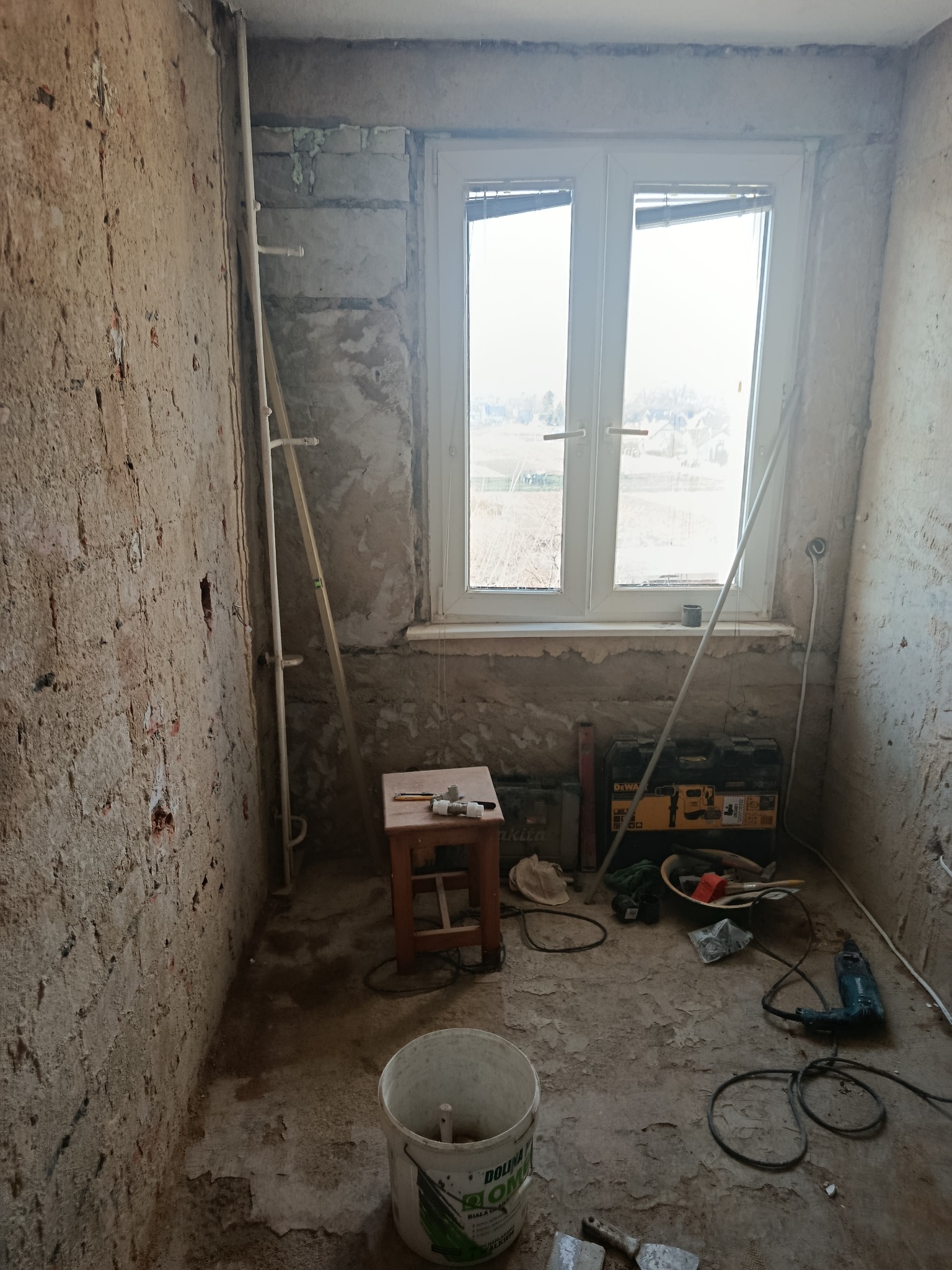 Wnętrze pokoju w trakcie remontu: surowe ściany, okno z widokiem na zewnątrz, narzędzia i akcesoria budowlane porozrzucane na podłodze. Widoczny etap przygotowania do prac wykończeniowych.