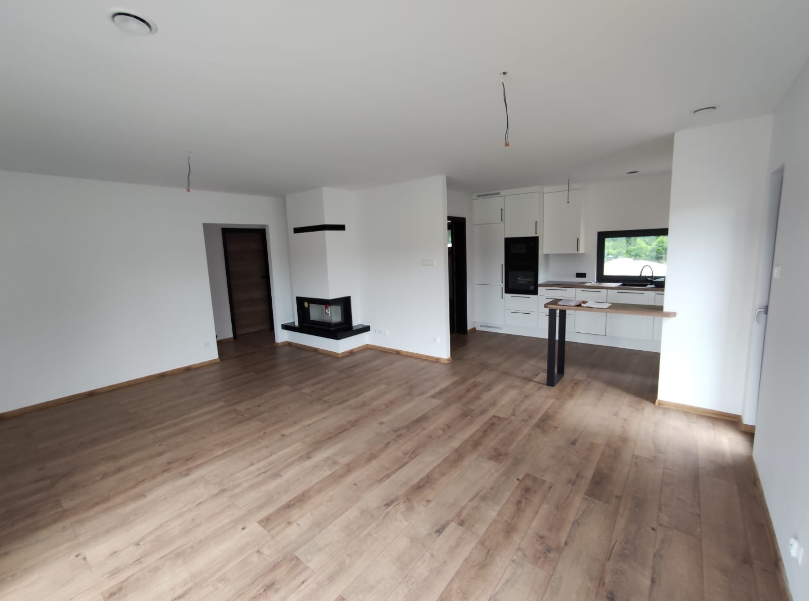Jasne, przestronne wnętrze po remoncie: drewnopodobna podłoga, białe ściany, nowoczesny kominek i kuchnia w tle. Widoczne niedokończone detale.