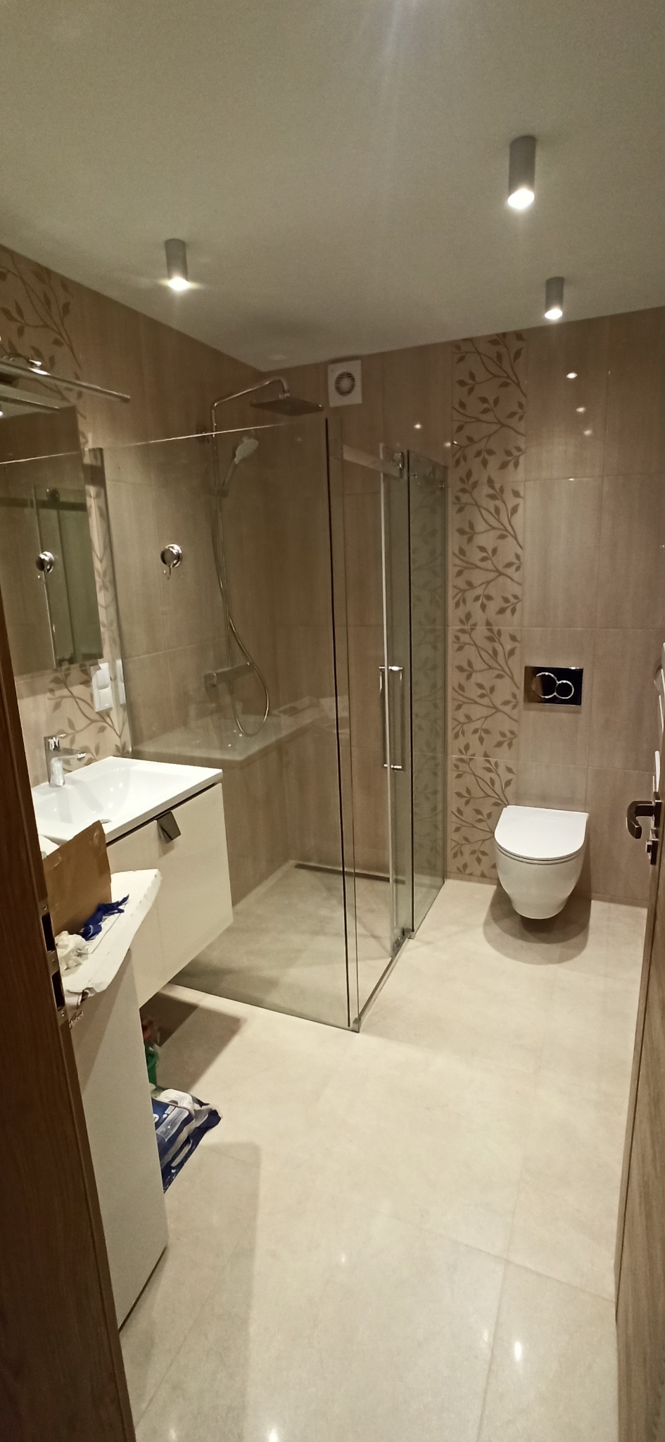 Jasna łazienka po remoncie z kabiną prysznicową, umywalką z szafką i toaletą. Na ścianach beżowe płytki z motywem roślinnym. Widoczne oświetlenie sufitowe.