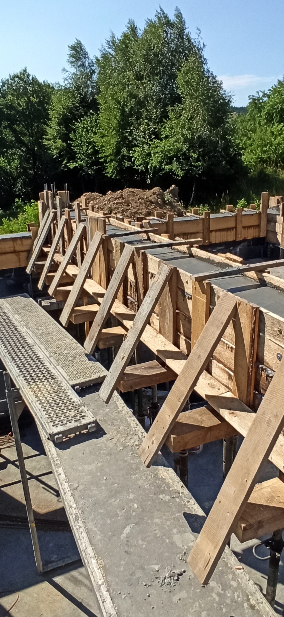Szczegółowe ujęcie drewnianego szalunku fundamentów z widocznym zbrojeniem, przygotowanego do zalania betonem na tle zieleni.