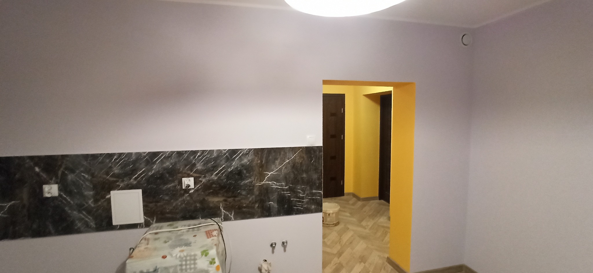 Wykończona kuchnia z szarymi ścianami, marmurowym panelem nad blatem i widokiem na żółty korytarz z drewnianą podłogą. Widoczne gniazdka i włączniki.