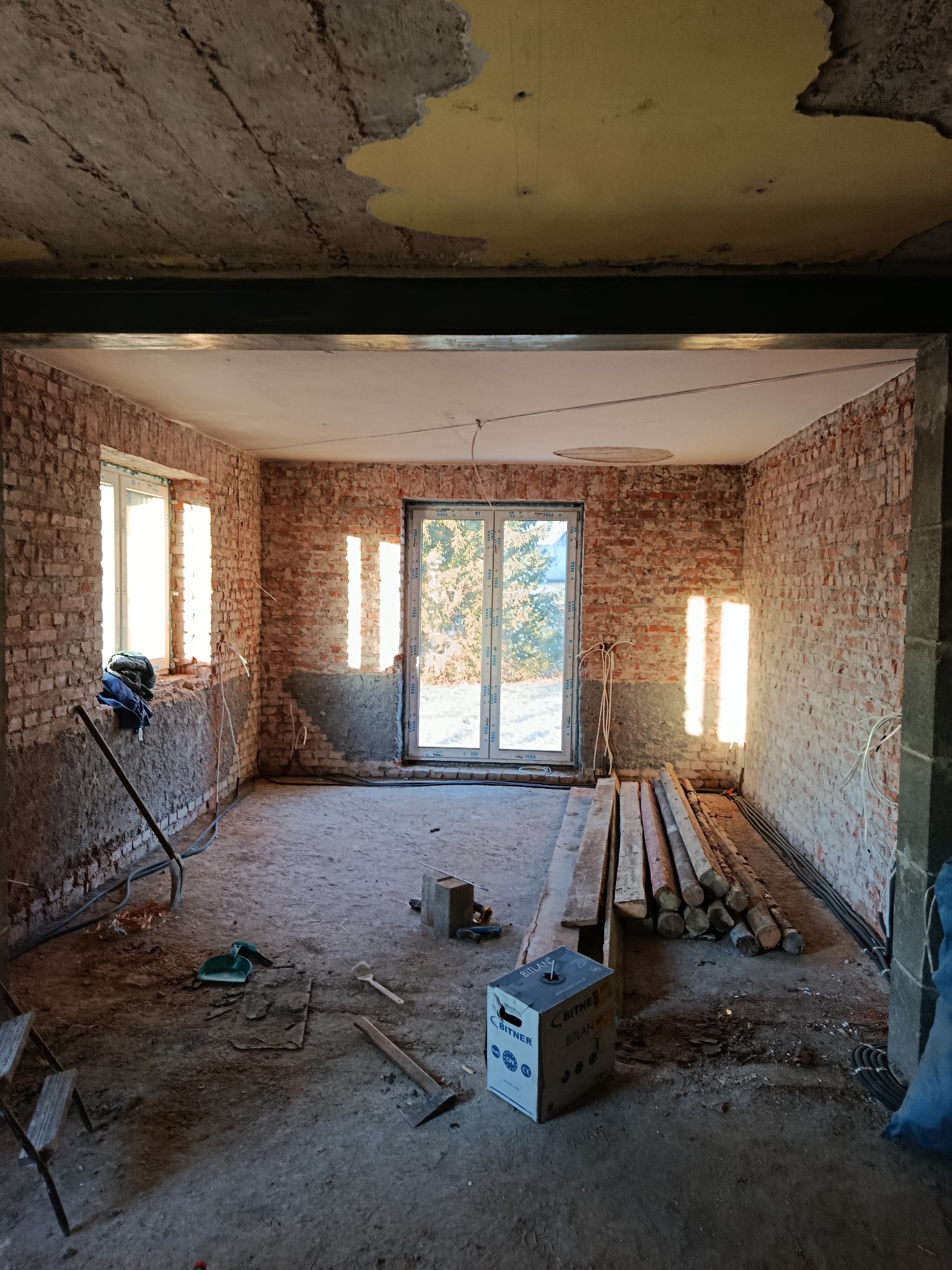 Pokój w trakcie remontu z odsłoniętą cegłą, widoczne przygotowania do dalszych prac wykończeniowych, materiały budowlane i narzędzia na podłodze, surowe ściany.