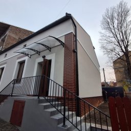 KOW-BUD - Dwukondygnacyjny dom z ocieploną elewacją w odcieniach szarości, z ceglanymi akcentami i zadaszeniem nad wejściem. Schody z czarną balustradą prowadzą do drzwi.