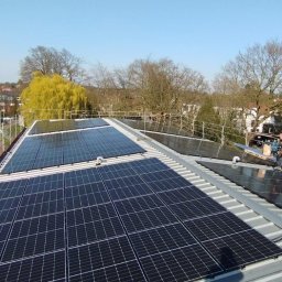 KOW-BUD - Instalacja paneli słonecznych na dachu budynku z widokiem na okolicę. Pracownik w trakcie montażu. Widoczne zabezpieczenia na dachu.