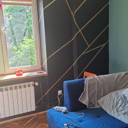 KOW-BUD - Salon z ciemną ścianą zdobioną złotymi, geometrycznymi wzorami. Na pierwszym planie niebieska sofa i drewniany stolik kawowy. Parkiet w jodełkę i okno z widokiem na zieleń.
