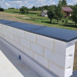 KOW-BUD - Świeżo wykończony mur z białych bloczków z czarnym, metalowym obiciem na płaskim dachu, na tle zieleni i zabudowań wiejskich w słoneczny dzień.