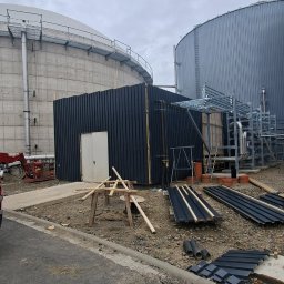 KOW-BUD - Budowa małego, czarnego budynku z blachy falistej na tle dwóch dużych zbiorników. Widoczne materiały budowlane i rusztowania. Pochmurne niebo.