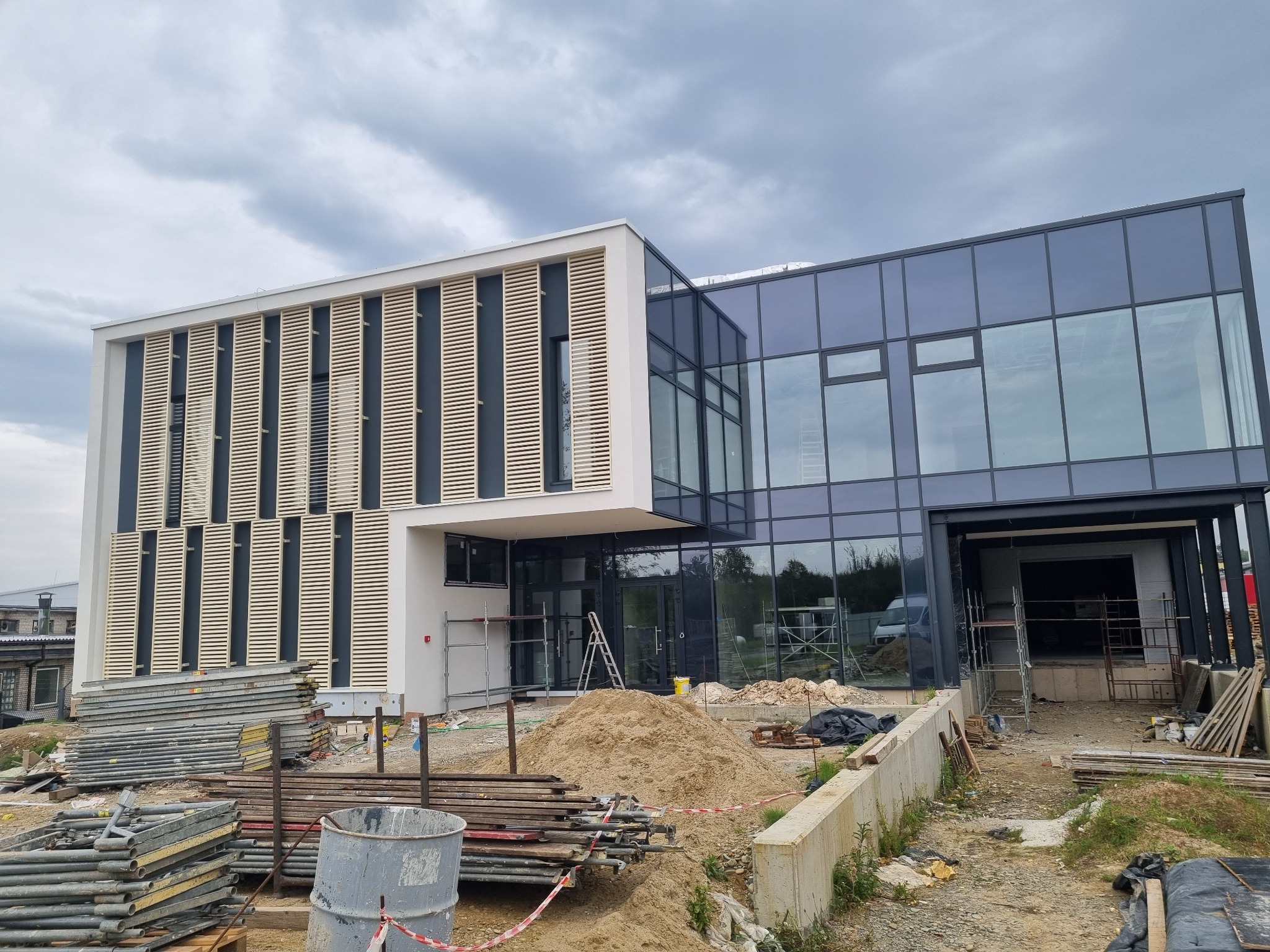 Nowoczesny budynek biurowy z elementami drewnianymi i szklaną fasadą w trakcie budowy, widoczne rusztowania i materiały budowlane.