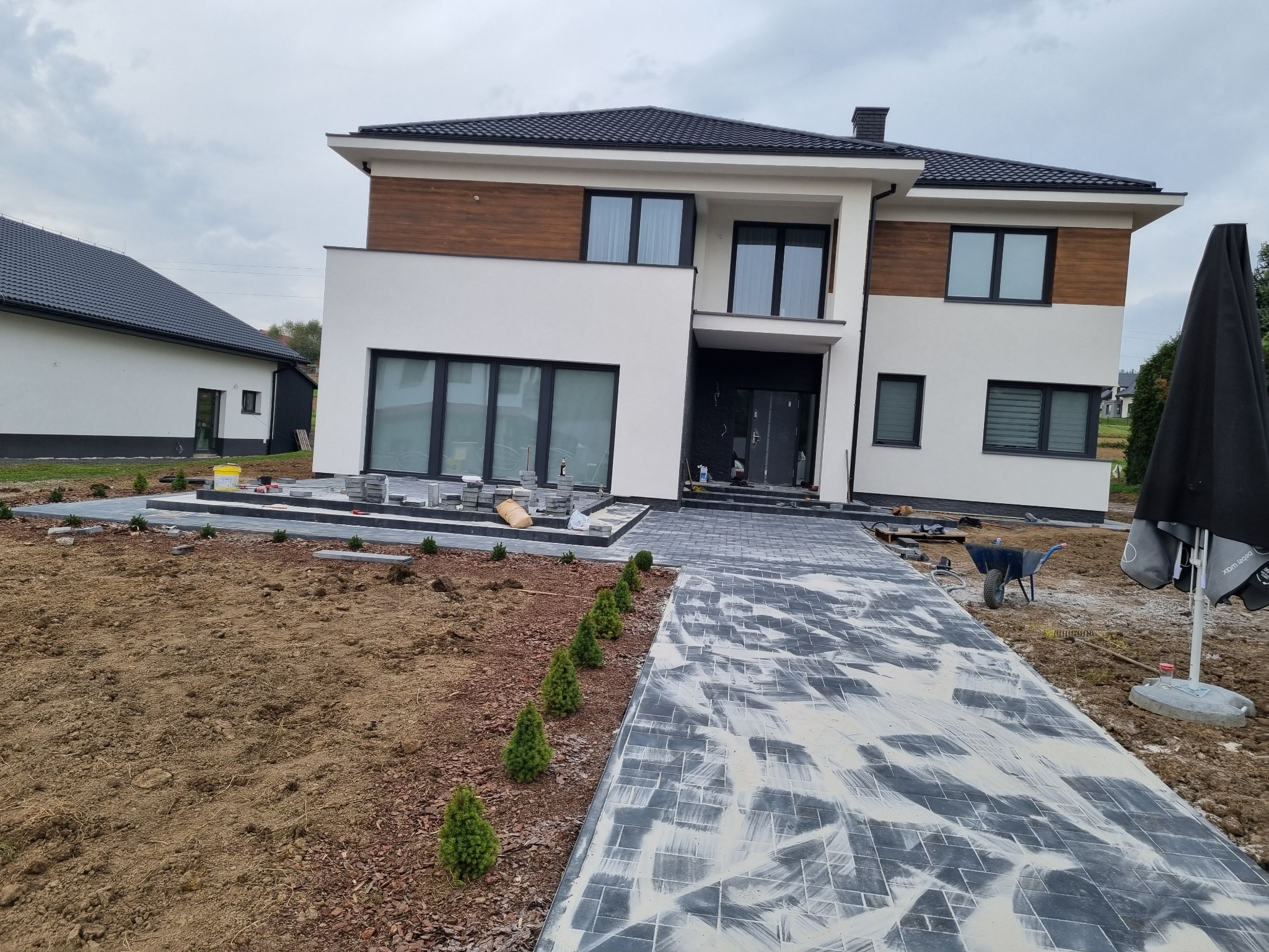 Nowoczesny dom z białymi ścianami i drewnianymi akcentami. Ułożona kostka brukowa prowadzi do wejścia. Widoczne prace wykończeniowe wokół budynku i świeżo posadzone rośliny.