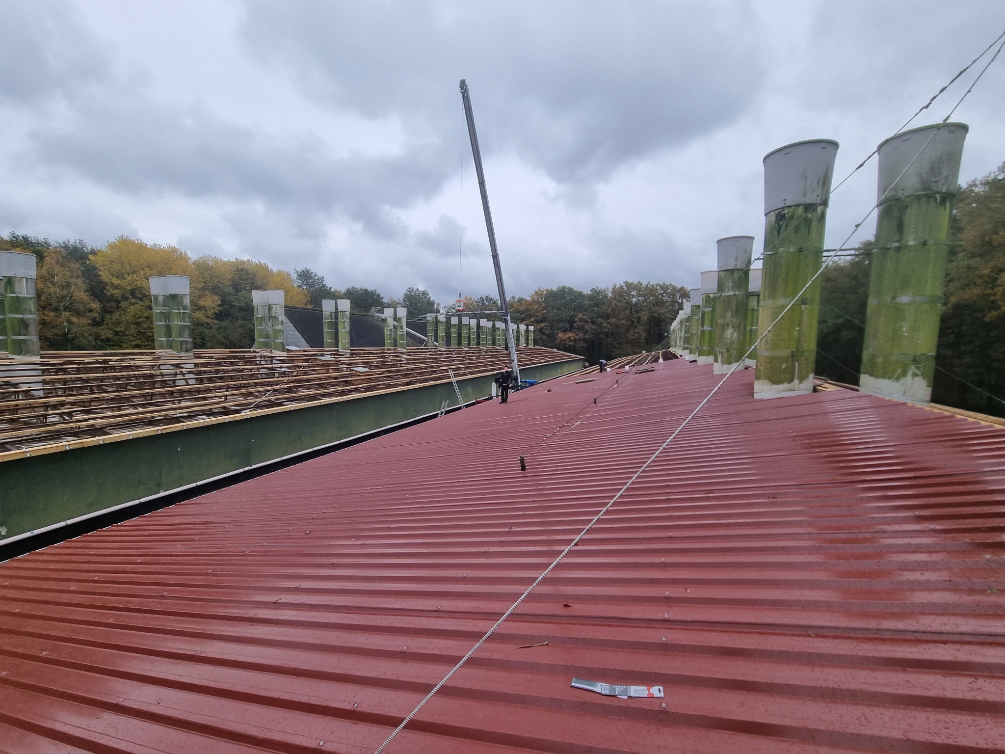 Czerwony dach z blachy trapezowej w trakcie montażu, widoczne kominy wentylacyjne z zielonym nalotem i konstrukcja drewniana dachu. Pochmurne niebo w tle.
