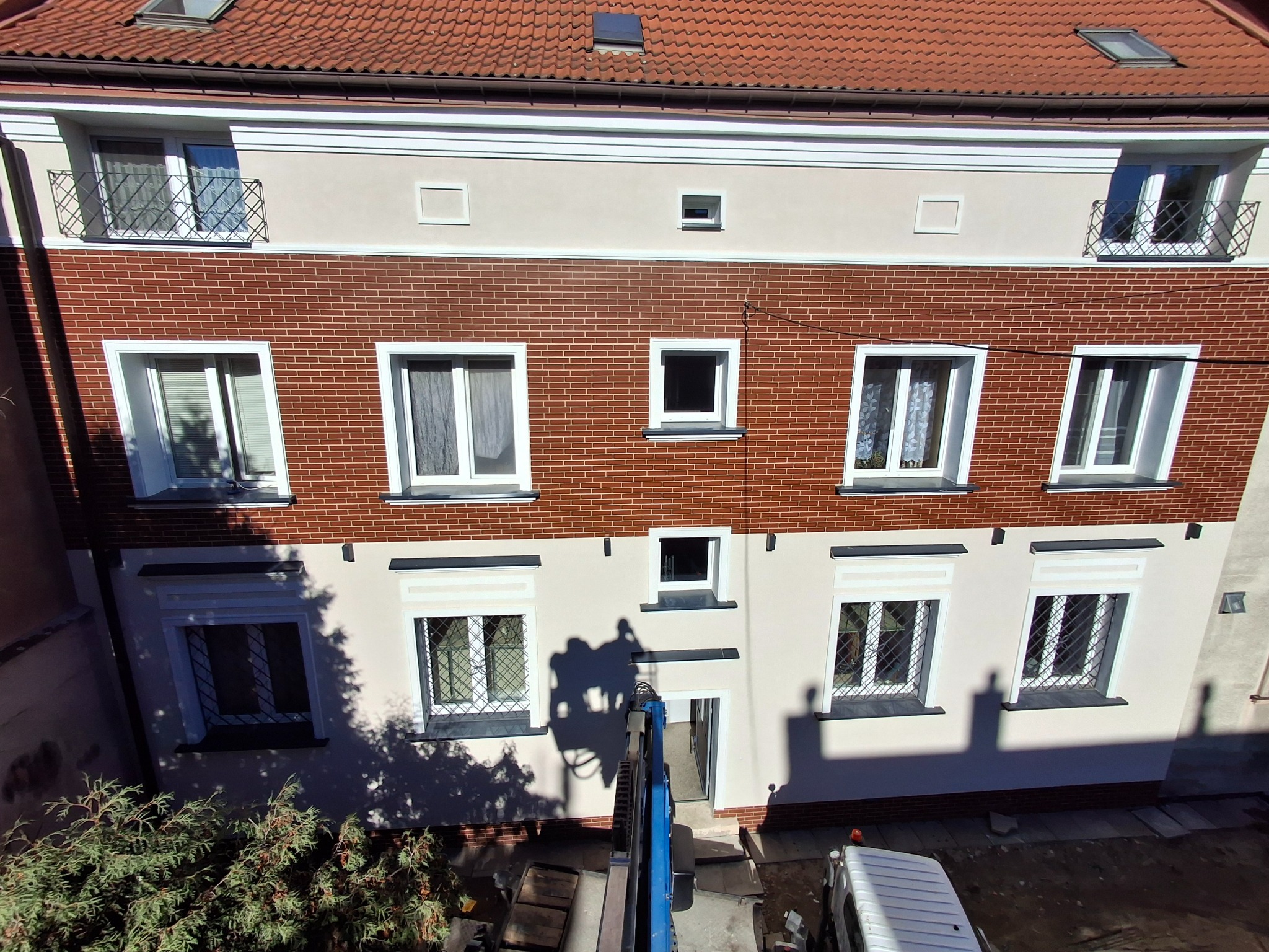 Odnowiona elewacja budynku z ceglaną wstawką, widziana z góry z platformy roboczej. Cień platformy na fasadzie. Białe ramy okienne i dekoracyjne elementy architektoniczne.