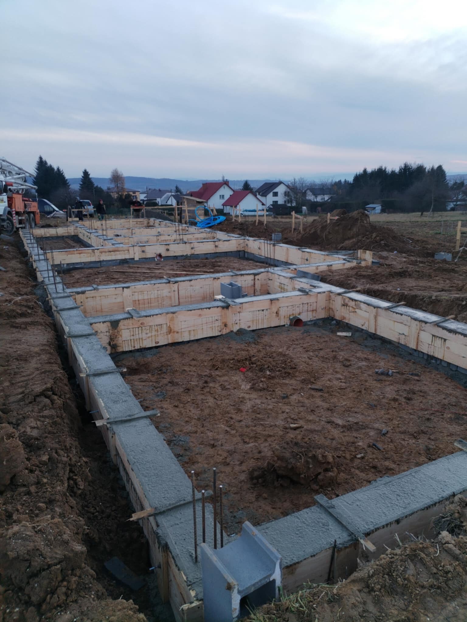 Zalane betonem szalunki fundamentów domu jednorodzinnego, widoczne zbrojenia i częściowo zasypana ziemią, w tle zabudowa jednorodzinna i pochmurne niebo.