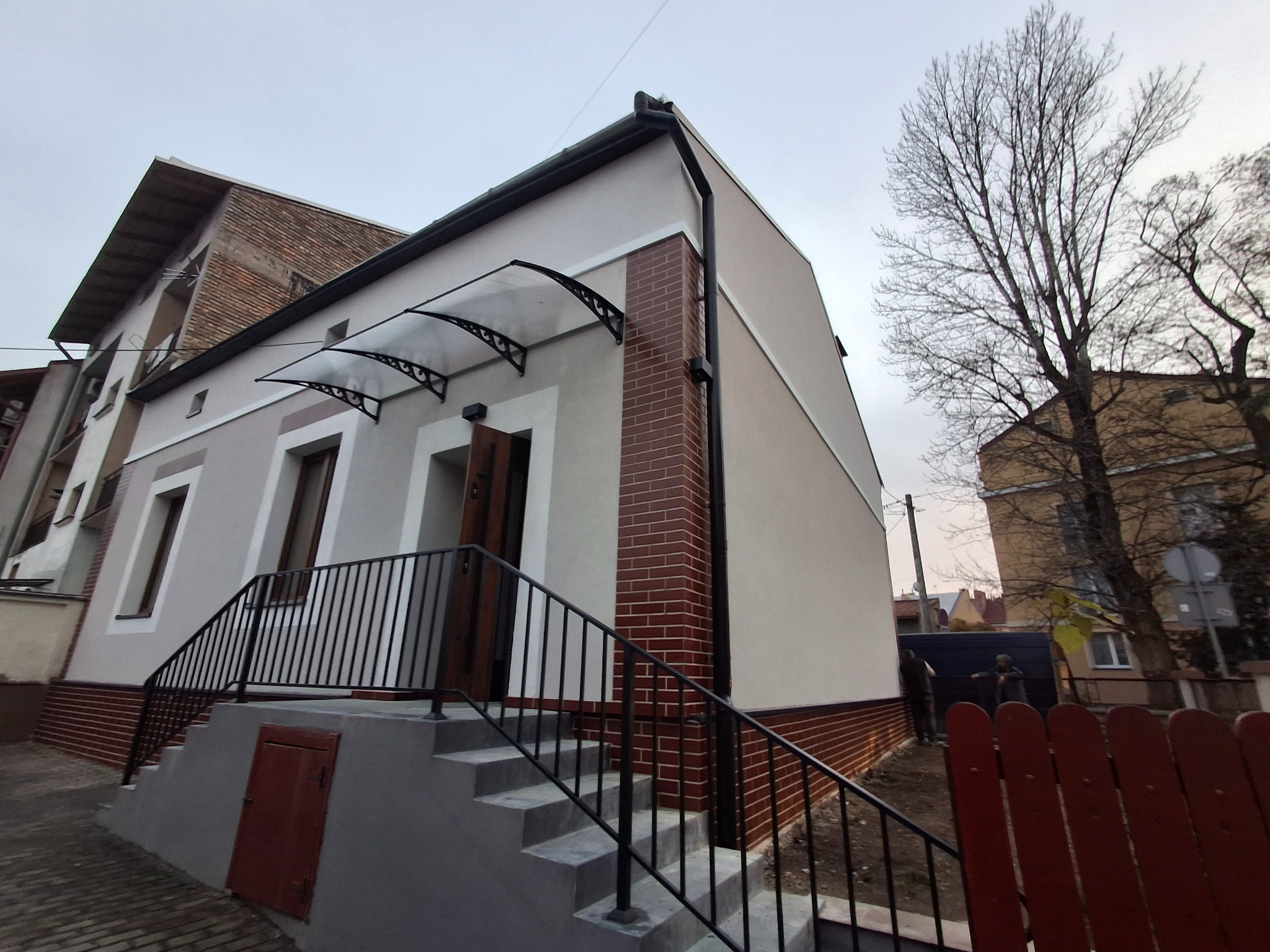 Dwukondygnacyjny dom z ocieploną elewacją w odcieniach szarości, z ceglanymi akcentami i zadaszeniem nad wejściem. Schody z czarną balustradą prowadzą do drzwi.