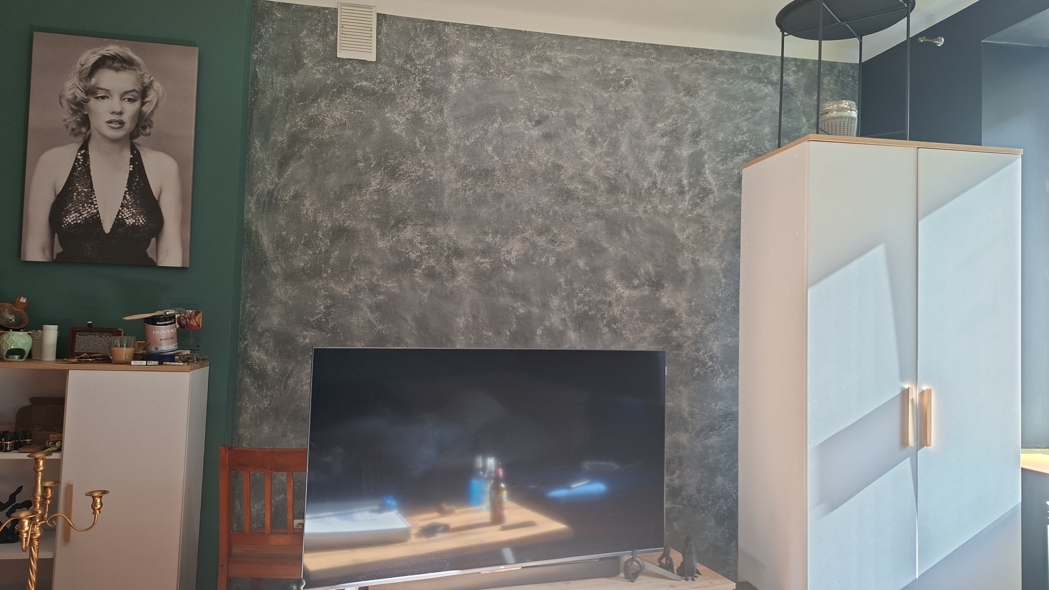 Szara ściana z efektem dekoracyjnym, obraz Marilyn Monroe na zielonej ścianie, biała szafka i telewizor. Widoczne elementy wystroju wnętrza.