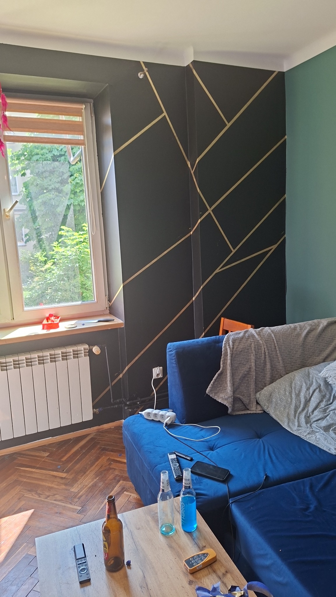 Salon z ciemną ścianą zdobioną złotymi, geometrycznymi wzorami. Na pierwszym planie niebieska sofa i drewniany stolik kawowy. Parkiet w jodełkę i okno z widokiem na zieleń.