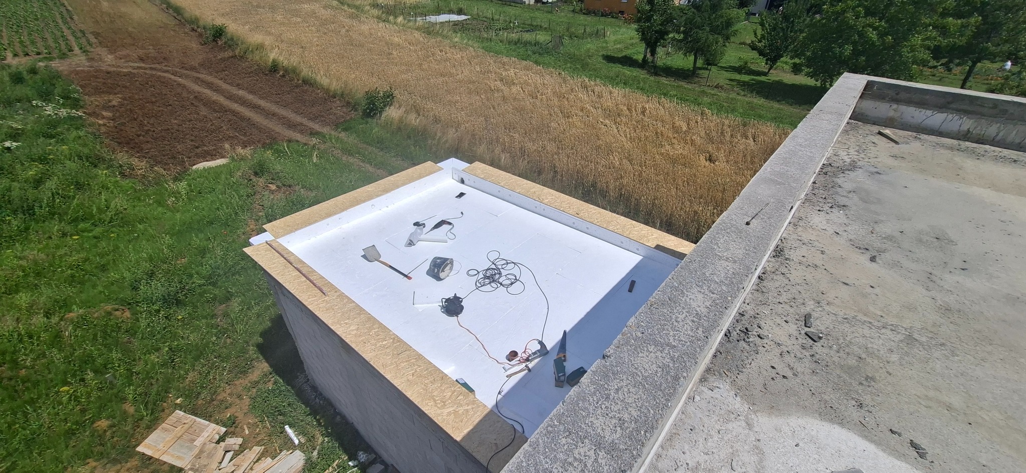 Izolacja dachu płaskiego styropianem w Jaśle. Widok z góry na białe płyty izolacyjne, narzędzia pracy i otaczające pola uprawne. Prace budowlane w toku.