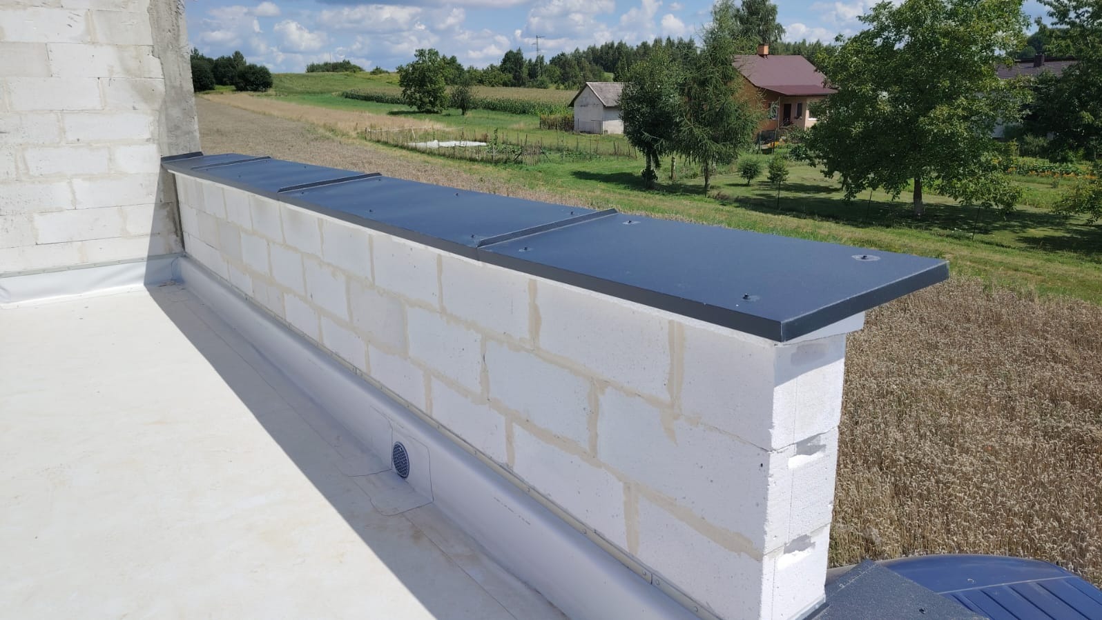 Świeżo wykończony mur z białych bloczków z czarnym, metalowym obiciem na płaskim dachu, na tle zieleni i zabudowań wiejskich w słoneczny dzień.