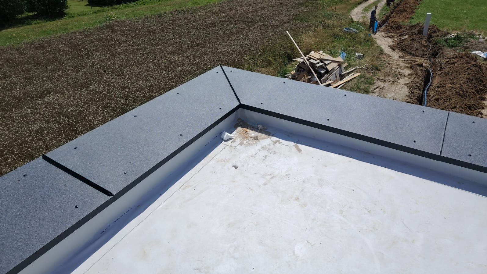 Wykończony fragment dachu płaskiego z szarą, matową obróbką blacharską, widok z góry na białą membranę dachową i otaczające pole. Prace dekarskie.