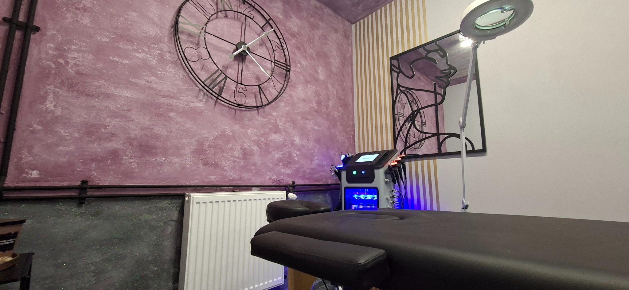 Klimatyczne wnętrze gabinetu kosmetycznego z łóżkiem zabiegowym, lampą powiększającą i nowoczesnym urządzeniem do zabiegów na tle ściany z dekoracyjnym zegarem.