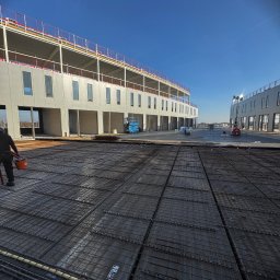 Krzysztof Wielgat - Zbrojenie pod wylewkę na budowie, widoczne budynki w stanie surowym otwartym, błękitne niebo. Prace przygotowawcze do montażu okien i drzwi.