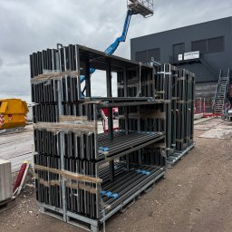 Krzysztof Wielgat - Sztaple aluminiowych ram okiennych na paletach, zabezpieczone folią, na placu budowy z podnośnikiem koszowym w tle. Widoczny budynek z napisem CantiVino.