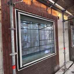 Krzysztof Wielgat - Montaż okna w budynku z cegły, widoczne rusztowanie. Okno z białą ramą i czarnym uszczelnieniem. Widok na budynek przez szybę. Prace budowlane w toku.