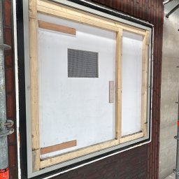 Krzysztof Wielgat - Montaż okna w murze z cegły, widoczne drewniane elementy konstrukcyjne i siatka zabezpieczająca. Etap prac wykończeniowych, rusztowanie w tle.