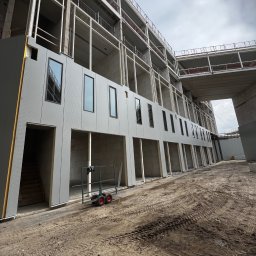 Krzysztof Wielgat - Budynek w trakcie montażu okien i elewacji, widoczne rusztowania i plac budowy. W tle pochmurne niebo.