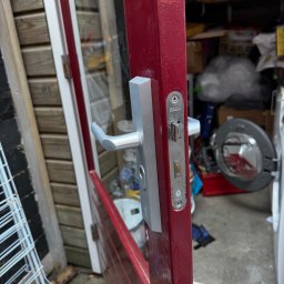 Krzysztof Wielgat - Częściowo otwarte, bordowe drzwi z srebrną klamką i zamkiem IVANA, prowadzące do zagraconego pomieszczenia z pralką. Widoczne ślady użytkowania na zamku.