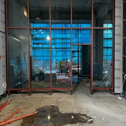 Krzysztof Wielgat - Montaż wielkoformatowych okien aluminiowych z widocznym rusztowaniem w tle. Prace w toku, widoczny pracownik na podnośniku. Surowy stan wykończenia.