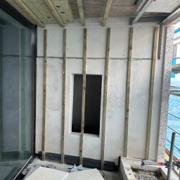 Krzysztof Wielgat - Montaż okna z drewnianą ramą i izolacją na balkonie w surowym stanie. Widoczne listwy montażowe i elementy wykończeniowe. Prace budowlane w toku.