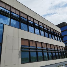 Krzysztof Wielgat - Elewacja budynku biurowego z rzędem okien z brązowymi roletami i odbiciem chmur. Widoczne elementy konstrukcyjne i sąsiedni budynek.