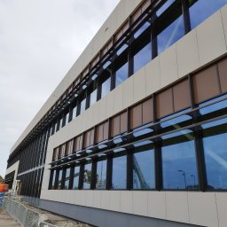 Krzysztof Wielgat - Elewacja budynku z oknami aluminiowymi i brązowymi roletami. W oknach odbija się niebo i dźwig budowlany. Widok z dołu, pochmurne niebo.