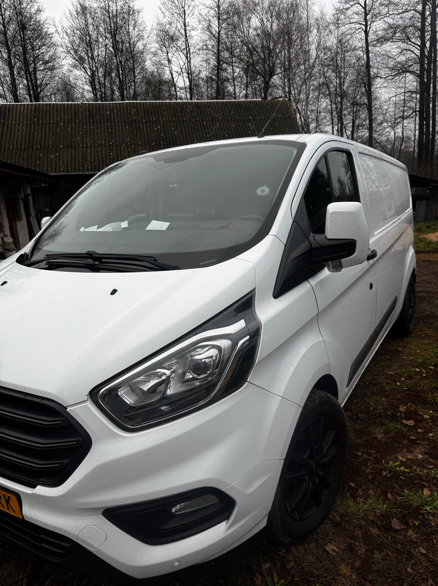 Biały samochód dostawczy na tle zarośniętego terenu i zadaszenia budynku. Widoczne detale karoserii, czarne felgi i fragment tablicy rejestracyjnej.