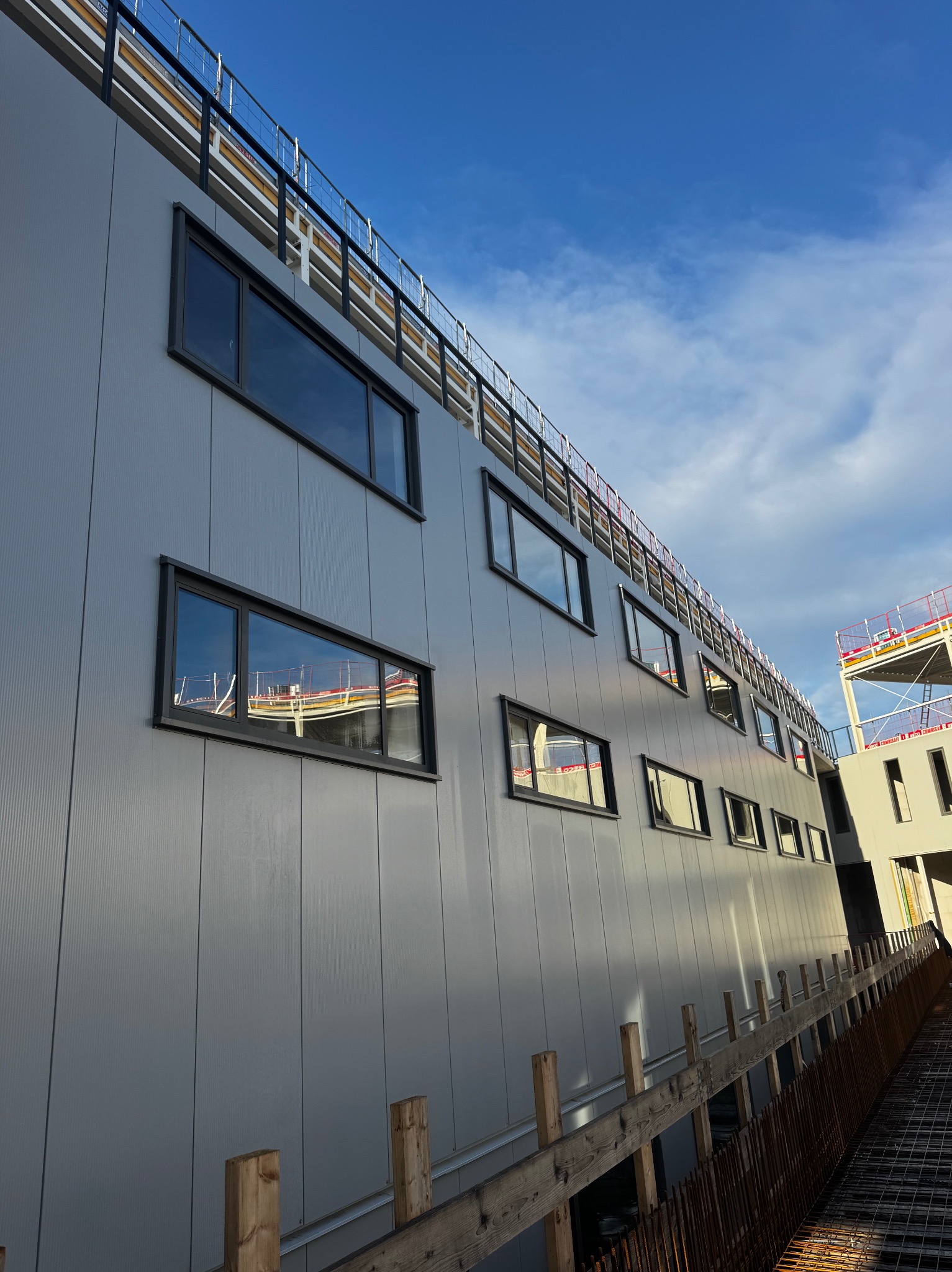 Nowoczesna fasada budynku z szarymi panelami i rzędem okien z czarnymi ramami, widoczna z dołu pod kątem, z elementami konstrukcyjnymi w tle i błękitnym niebem.