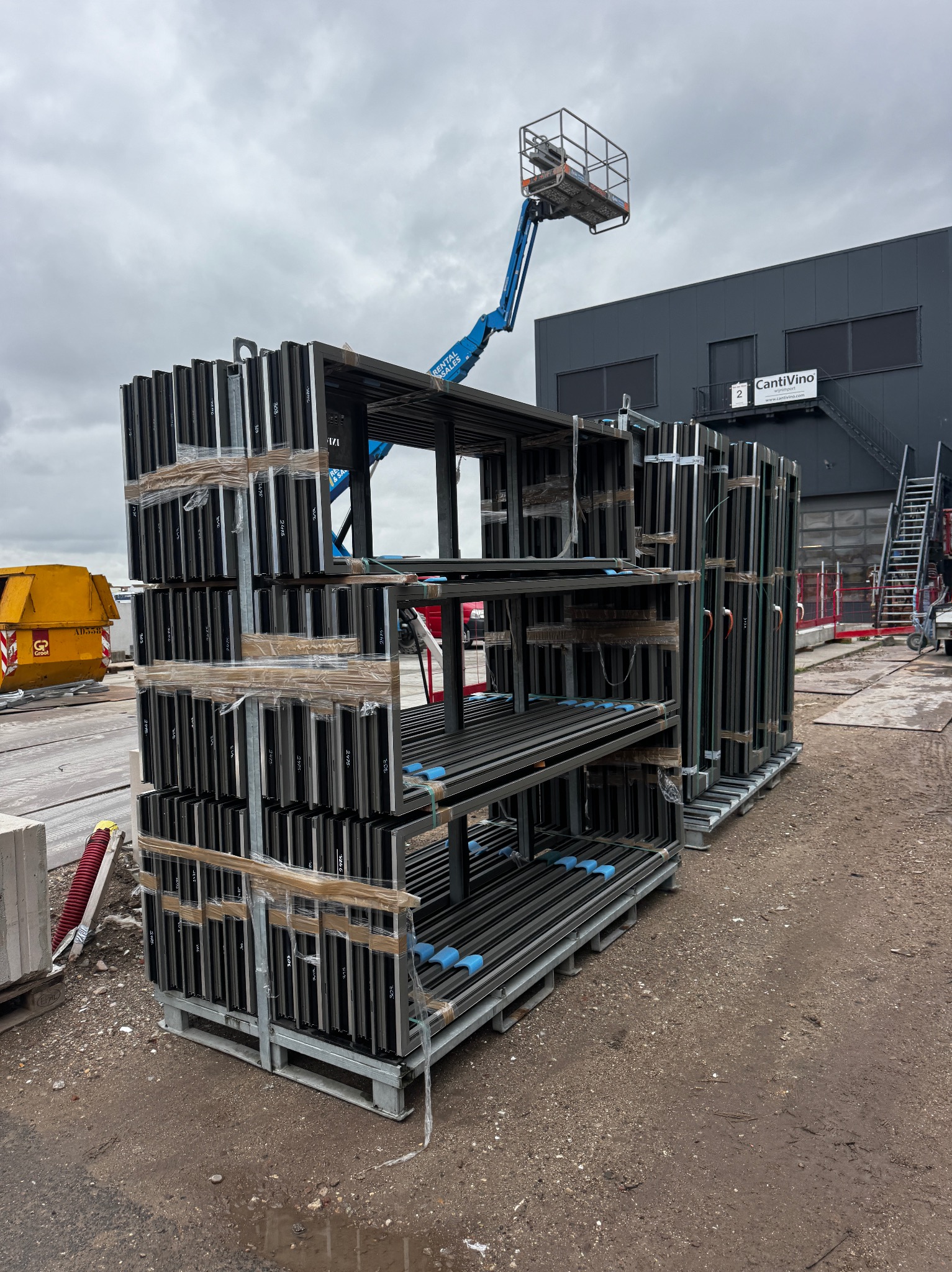 Sztaple aluminiowych ram okiennych na paletach, zabezpieczone folią, na placu budowy z podnośnikiem koszowym w tle. Widoczny budynek z napisem CantiVino.