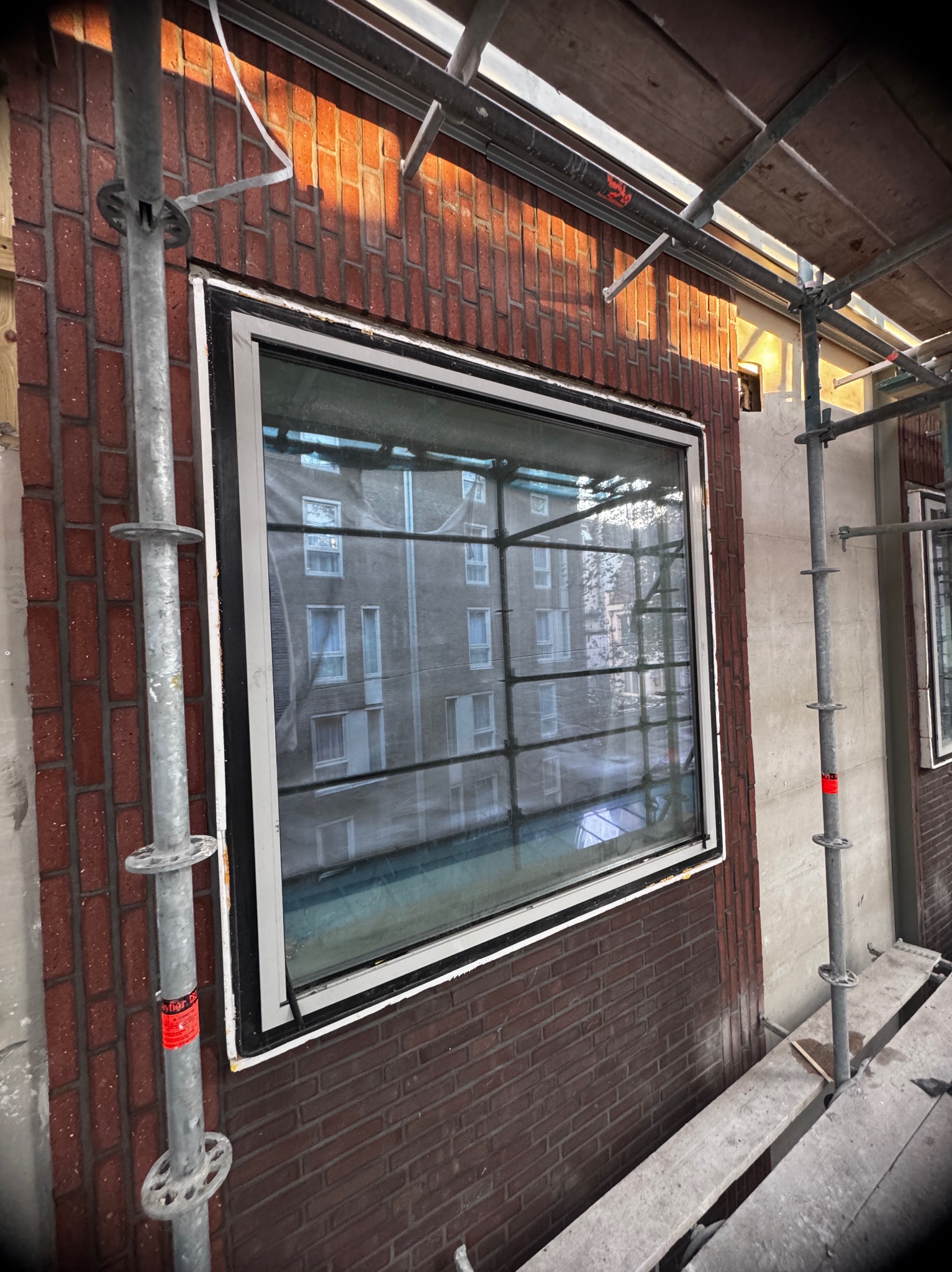 Montaż okna w budynku z cegły, widoczne rusztowanie. Okno z białą ramą i czarnym uszczelnieniem. Widok na budynek przez szybę. Prace budowlane w toku.