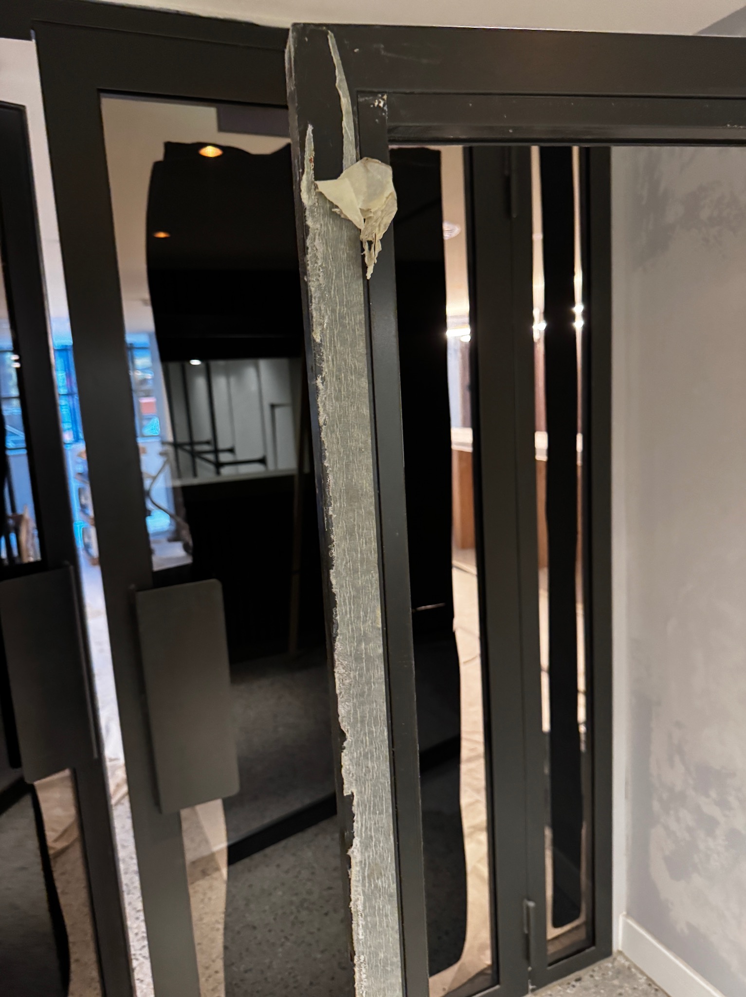 Uszkodzona, czarna rama drzwi z resztkami taśmy malarskiej. Widoczne wnętrze pomieszczenia przez szybę. Detal uszkodzenia ramy.