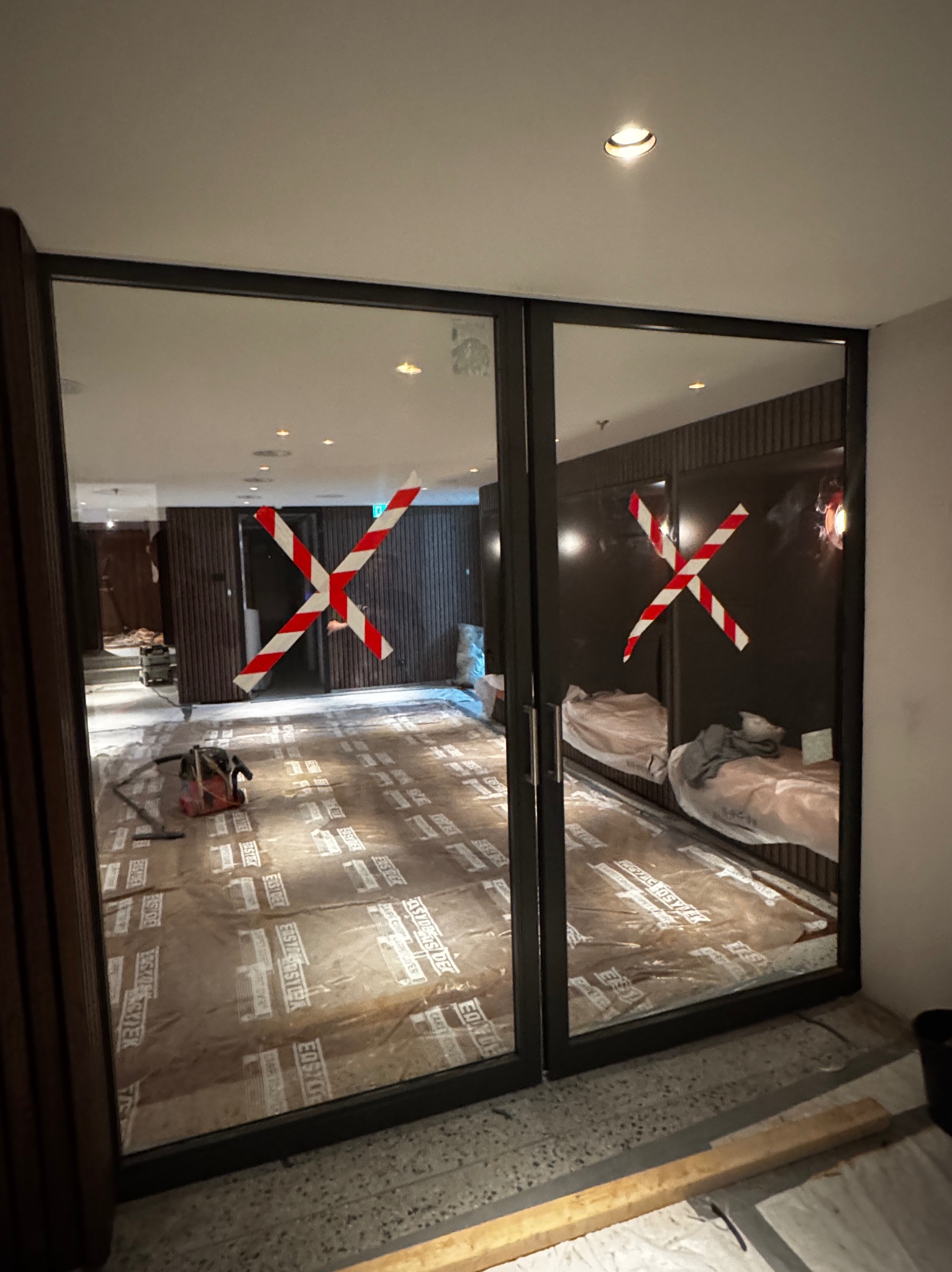 Ciemna rama przesuwnych drzwi z szybami, zabezpieczonymi taśmą ostrzegawczą, widoczna na tle remontowanego wnętrza z podłogą zabezpieczoną folią.