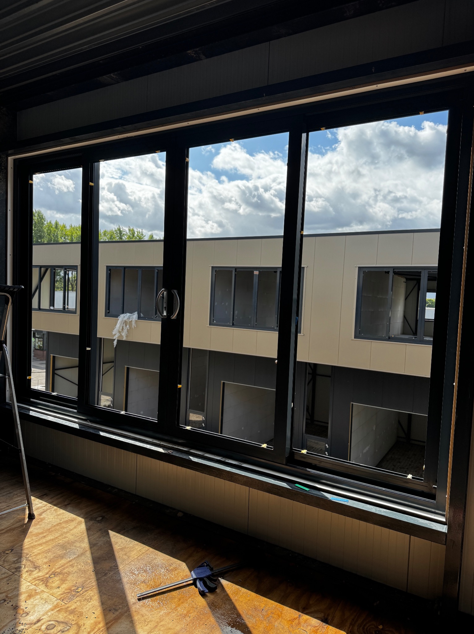 Nowe okno aluminiowe z widokiem na budynek w budowie. Widoczne narzędzia i drabina. Jasne światło słoneczne podkreśla detale konstrukcyjne.