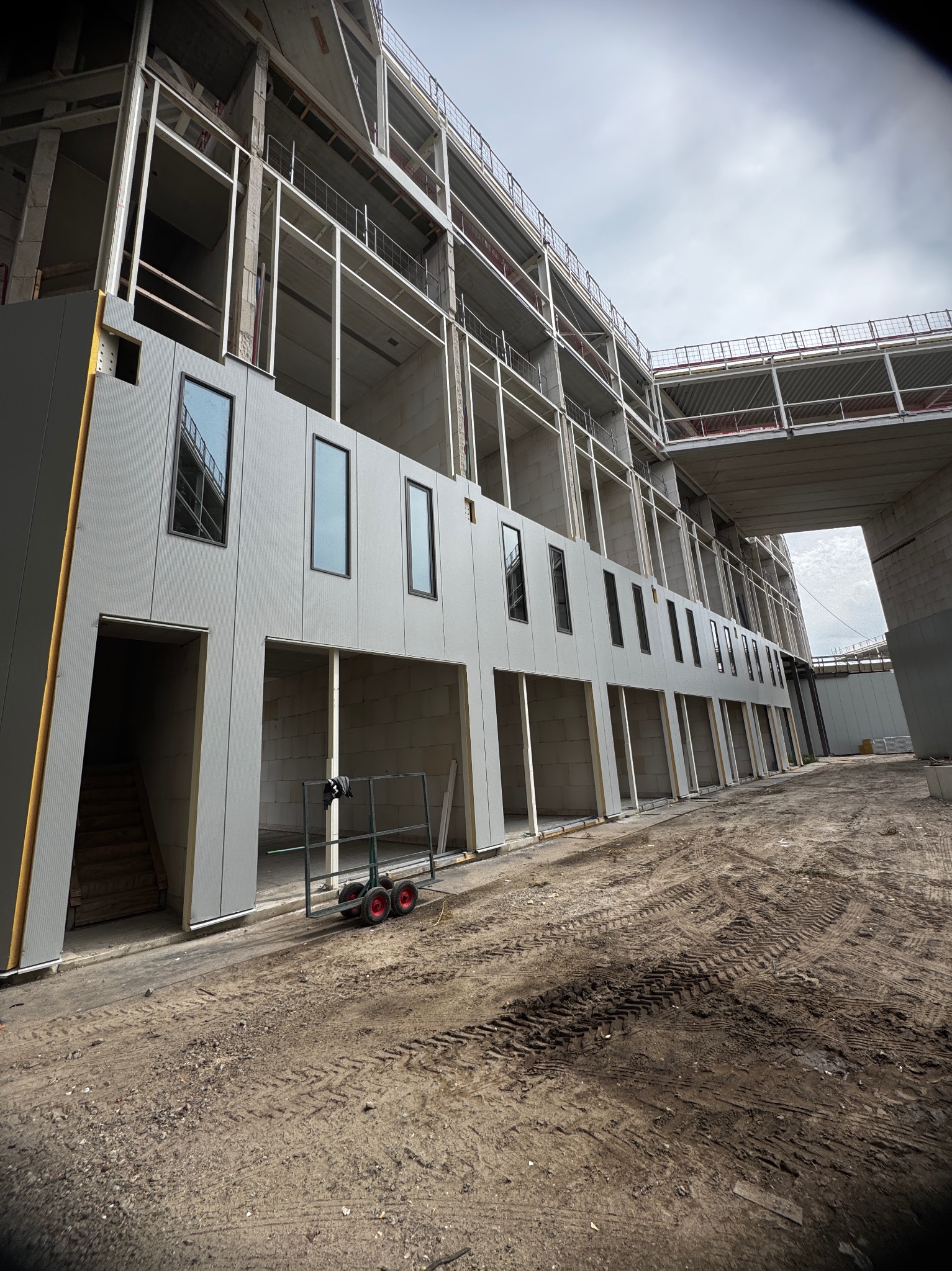 Budynek w trakcie montażu okien i elewacji, widoczne rusztowania i plac budowy. W tle pochmurne niebo.