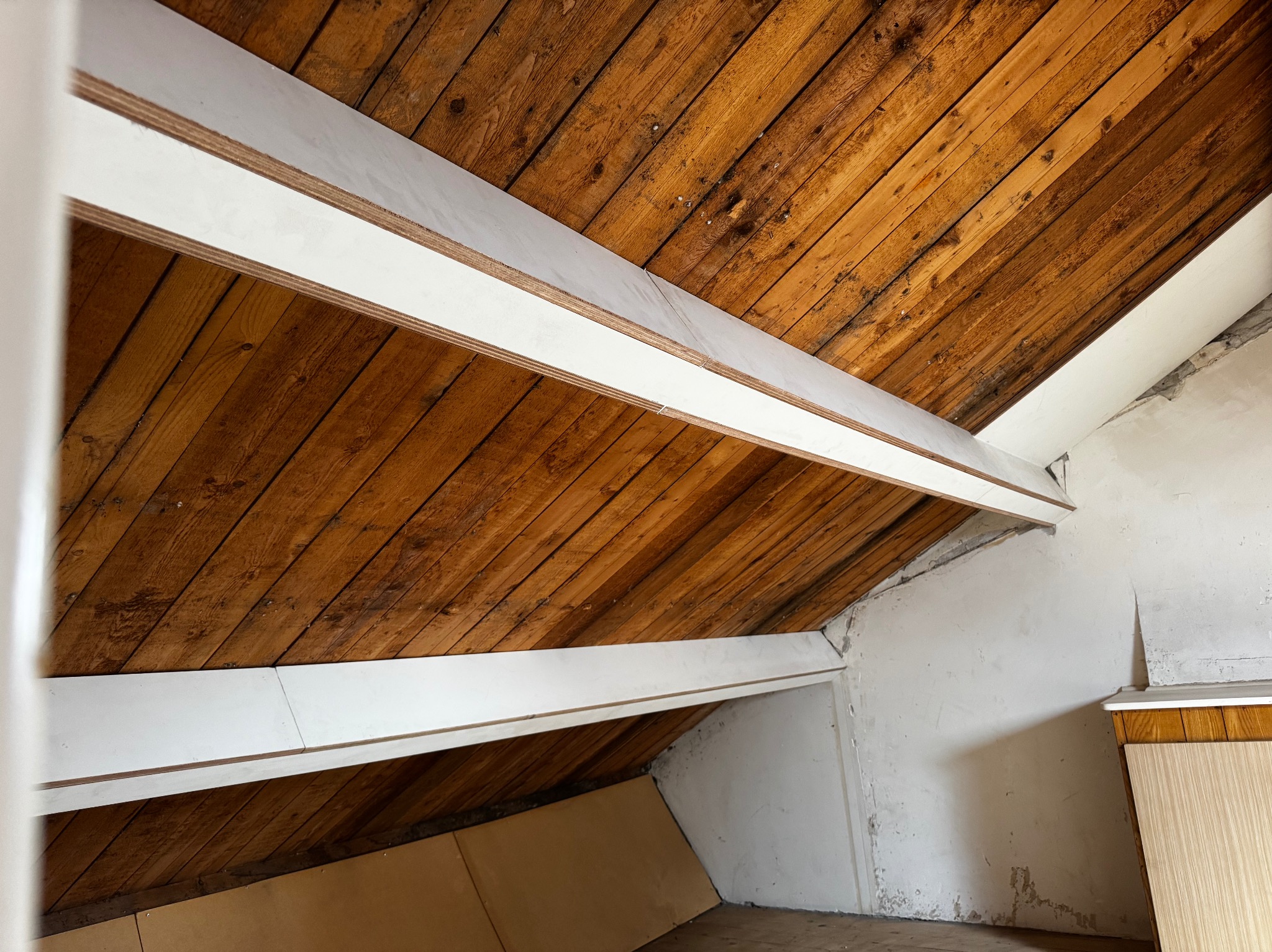 Skośny sufit z drewnianych desek, wzmocniony białymi belkami. Widoczne niedoskonałości ścian i podłogi, sugerujące remont lub adaptację poddasza.